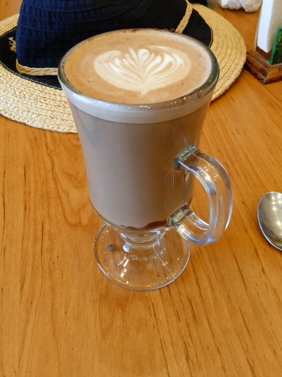 Capuchino con arte latte en taza de vidrio, junto a un sombrero sobre la mesa de madera